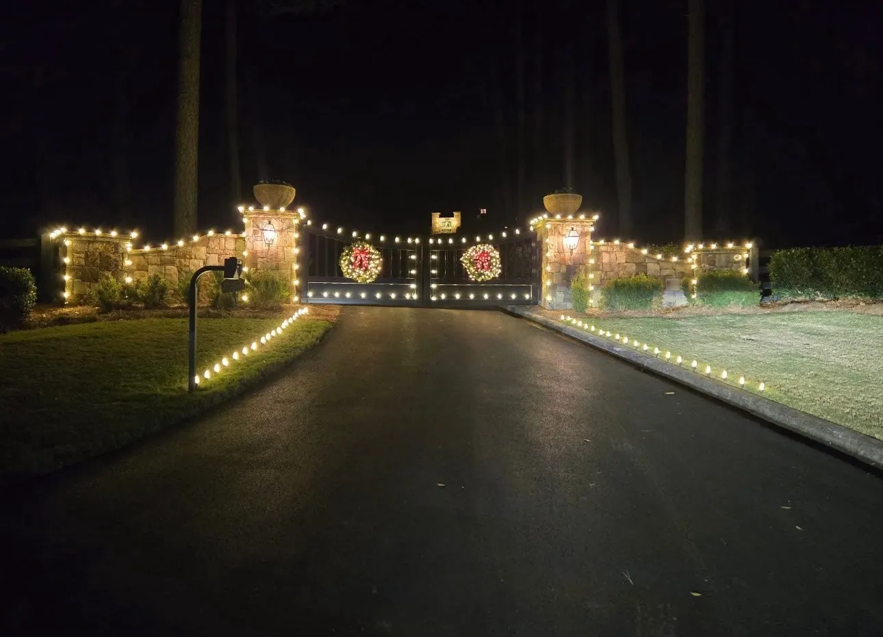 A stone entrance gate with warm white lights along the pillars and driveway edge, and two wreaths with red bows on the gate.