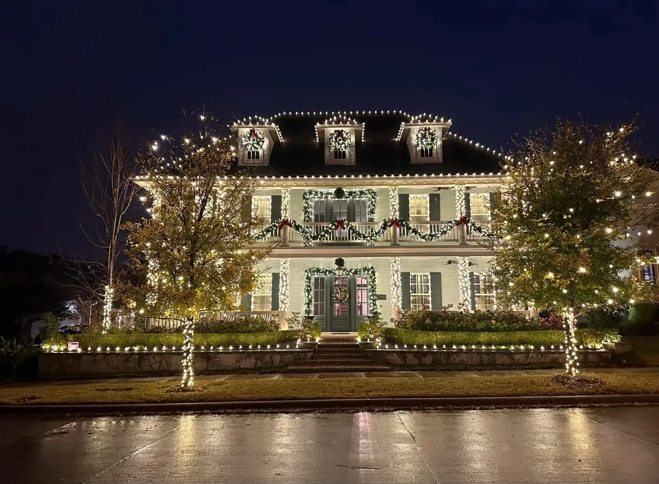 A large three-story home with roofline and dormer lighting, window wreaths, and garland with red bows along the balcony railings.