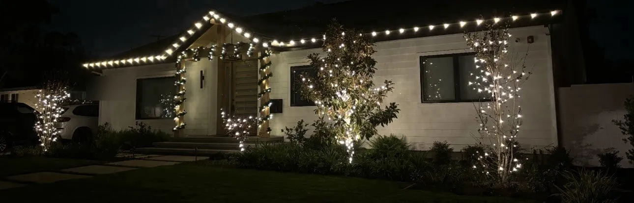 A single-story home with roofline lighting and two trees wrapped in warm white lights.