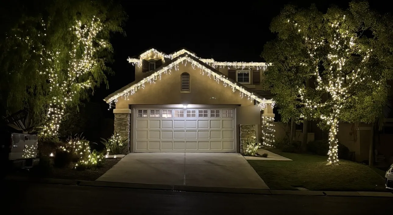 A two-story home with icicle-style roofline lighting and two willow trees wrapped in warm white lights.