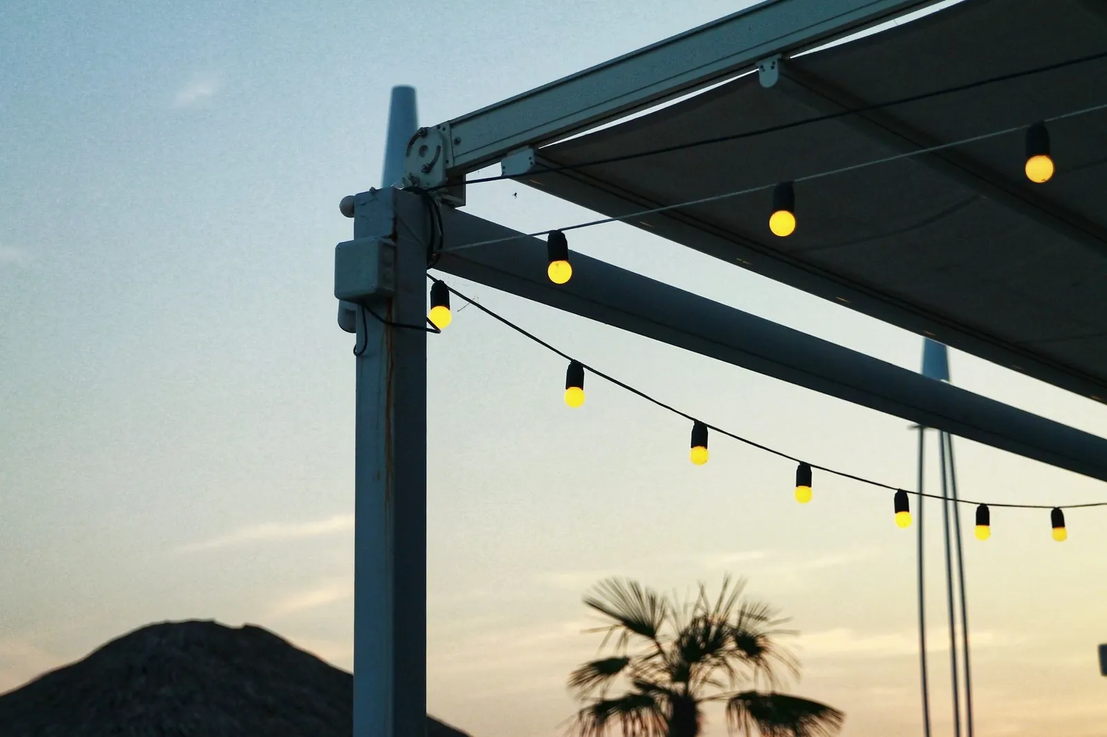Amber string-light bulbs strung from a white pergola frame at sunset, with palm fronds behind.