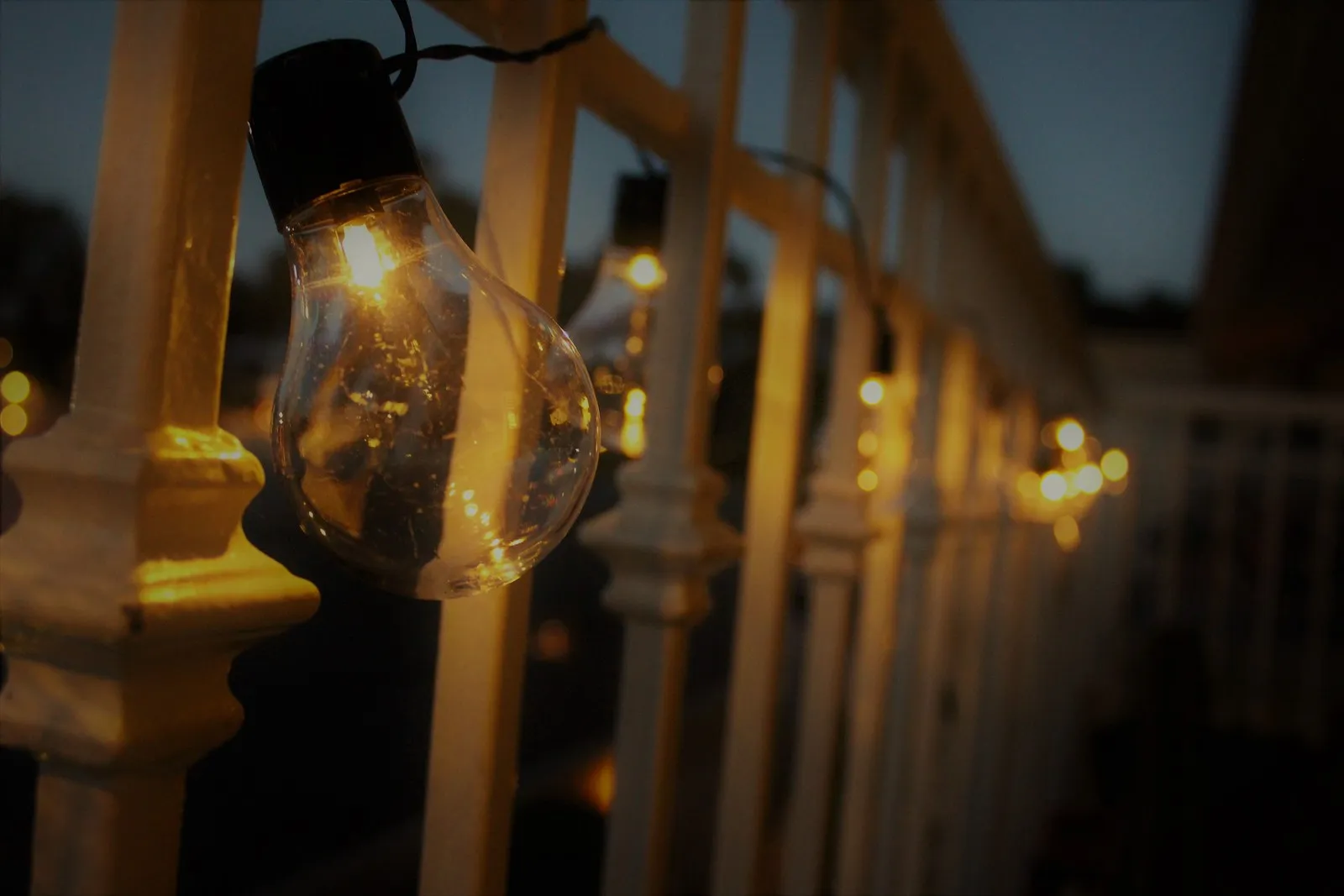 A close-up of one warm string-light bulb along a white railing at dusk.