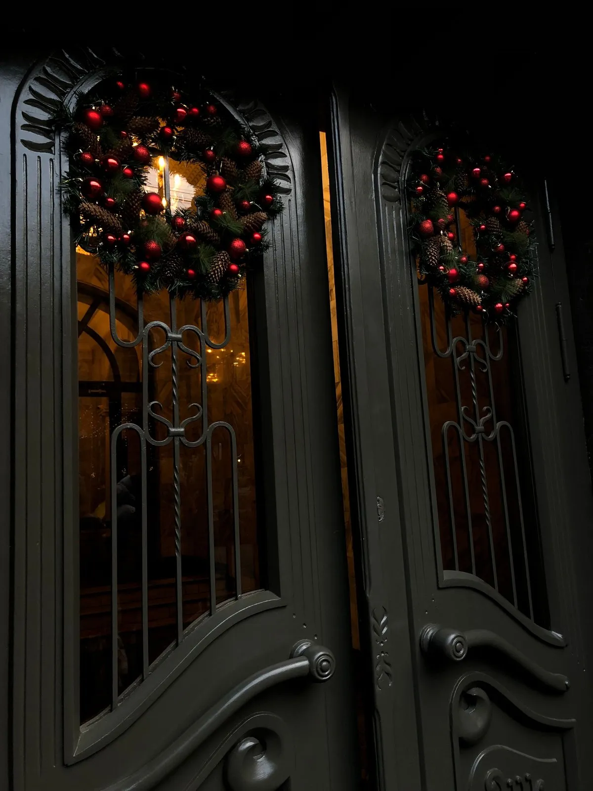 Dark double doors with evergreen wreaths dressed in red ornaments, warm light glowing through the glass.